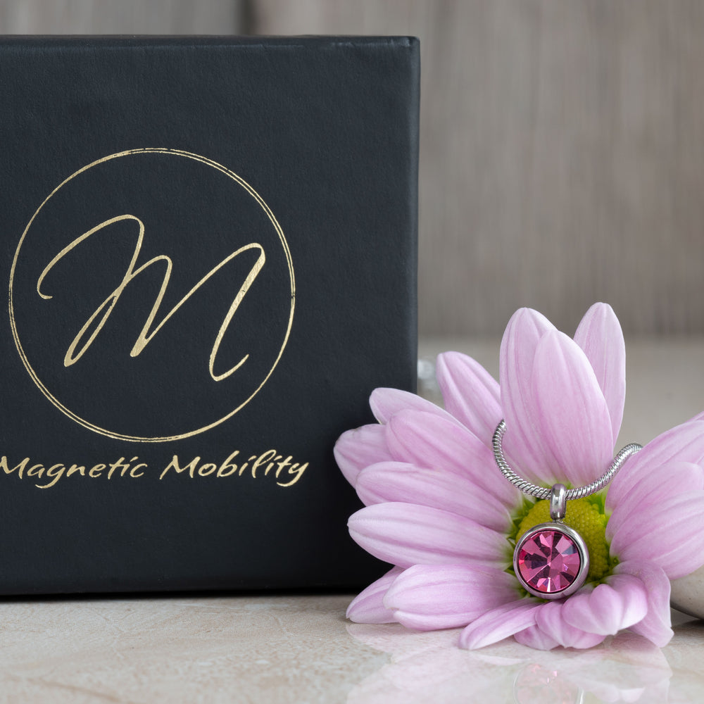 Close-up view of a pink tourmaline magnetic necklace, highlighting the sparkling pink gemstone set in a sleek, silver-toned pendant. The necklace is displayed alongside a pink flower and the Magnetic Mobility gift box.