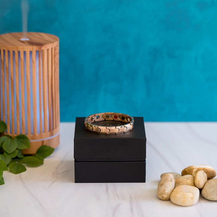 The image displays a rose gold Rowan Dawn men's bracelet. Its design consists of linked pieces with a matte inlay and shiny edge stripe for texture contrast. It features a secure clasp for ease of use. This accessory serves as a style statement and provides relief from conditions like arthritis, back pain, and migraines