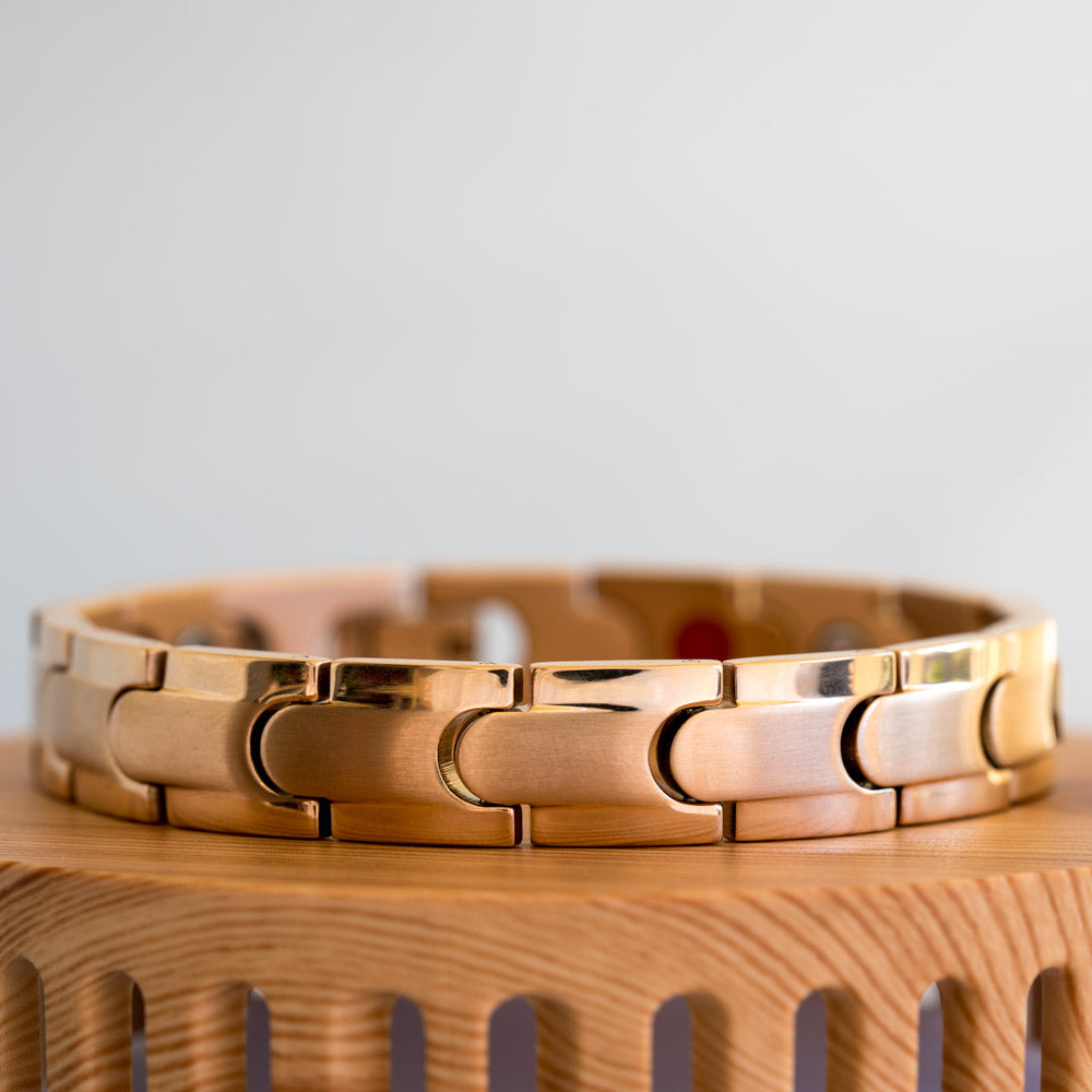Close-up view of a Rowan Dawn men's bracelet, illustrating the attention to detail in the construction. This rose gold-toned, stainless steel bracelet exhibits a texture contrast with a matte inlay and a shiny metallic stripe encircling the edge of each link. The secure clasp, integral to the design, ensures a comfortable fit. The image emphasizes the bracelet's dual-purpose as a stylish accessory and a potential aid for conditions such as arthritis, back pain, and migraines.