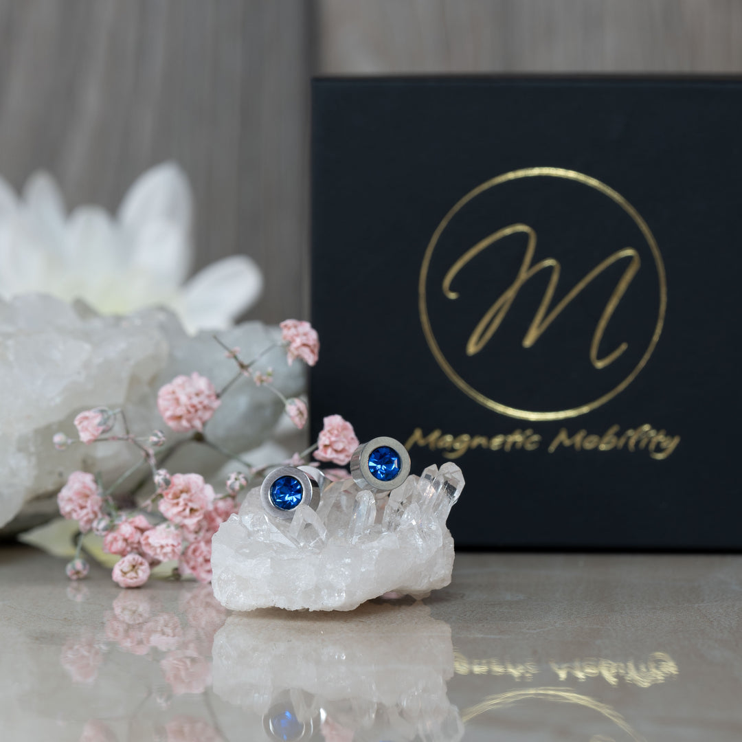 September Birthstone magnetic earrings with radiant Swarovski Blue Sapphire, elegantly displayed on a white crystal with pink flowers and a black Magnetic Mobility gift box as a backdrop. Perfect for adding a touch of elegance to September birthdays.