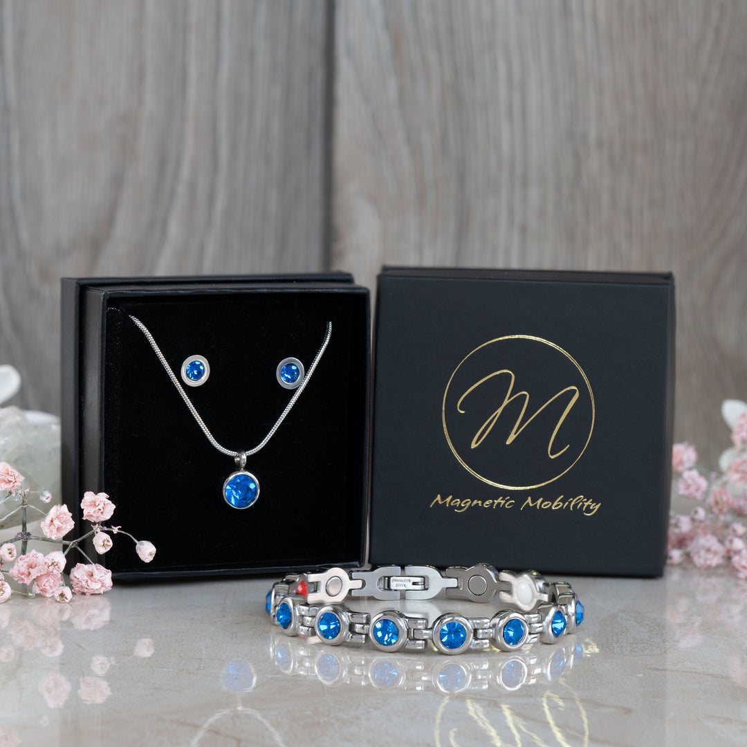 Complete view of the September Birthstone magnetic gift set including a necklace, earrings, and bracelet adorned with Swarovski Blue Sapphire crystals, arranged stylishly with pink flowers and a black gift box. Ideal for September birthday gifts, combining elegance and health benefits.