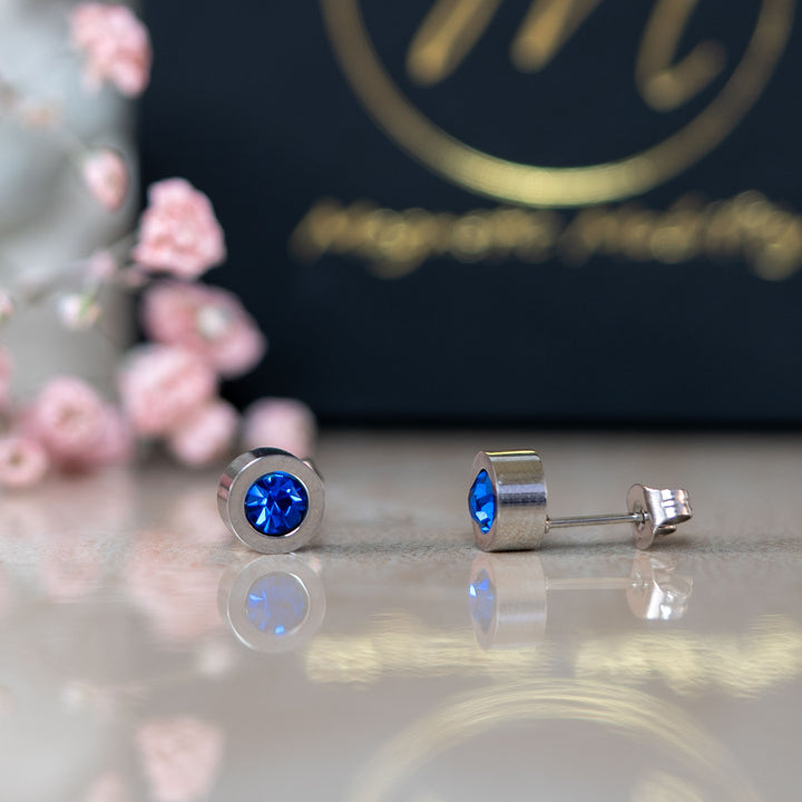 Close-up view of the September Birthstone magnetic earrings featuring striking Swarovski Blue Sapphire crystals, set against a backdrop of pink flowers and a black gift box. Ideal for celebrating September birthdays with style and wellness.