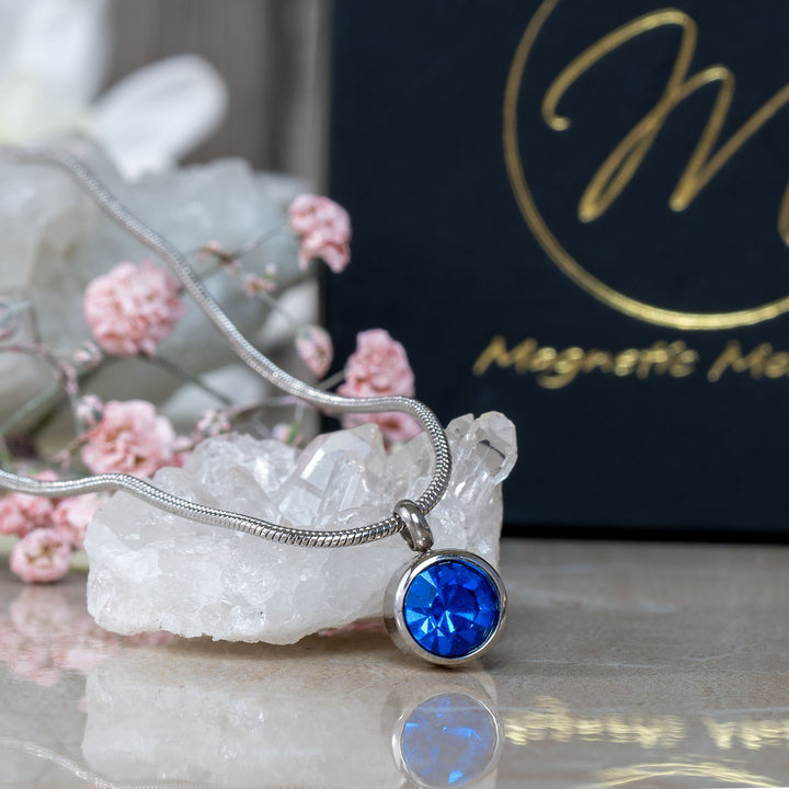 The September Birthstone magnetic necklace featuring a striking Swarovski Blue Sapphire pendant, beautifully displayed on a white crystal with pink flowers and a black gift box in the background. Ideal for adding a touch of elegance and wellness to September birthdays.