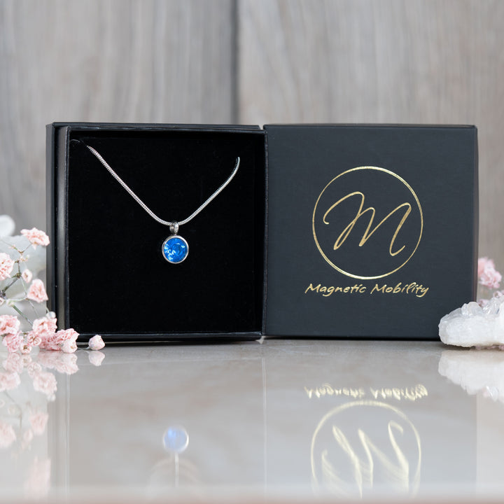 Elegant September Birthstone magnetic necklace featuring a vibrant Swarovski Blue Sapphire pendant, presented in a luxurious black box against a backdrop of pink flowers and white crystals. Perfect for adding sophistication to September birthday celebrations.