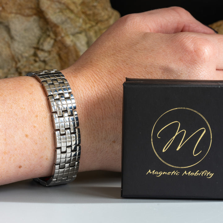 Wearer showcasing Magnetic Mobility's silver arthritis relief bracelet on the wrist, positioned next to a branded black box, with a natural stone background.