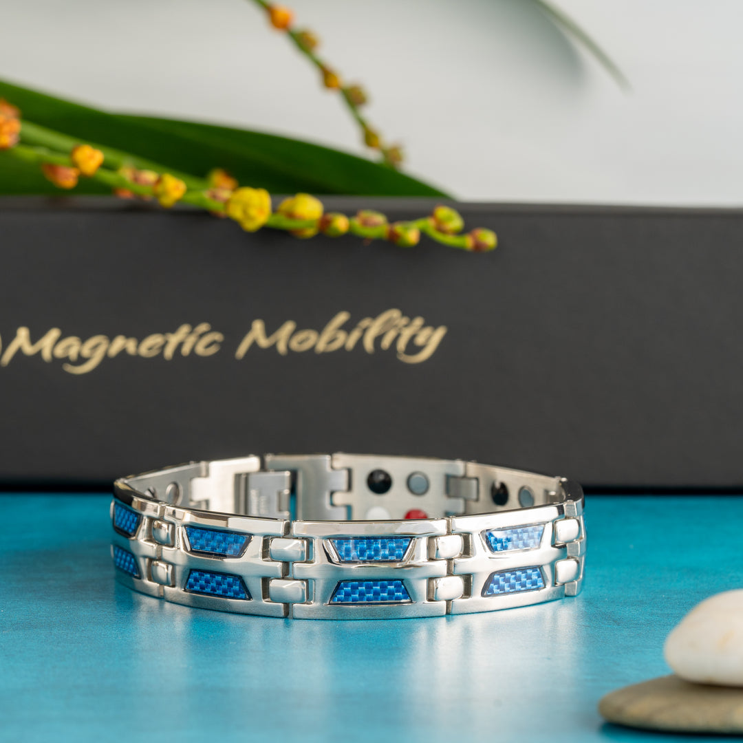 A Yarrow Star 4in1 Magnetic Bracelet from Magnetic Mobility rests on a blue surface, showcasing its unique silver design with blue carbon fiber inlays and therapeutic magnets visible on the inner side. The Magnetic Mobility's elegant black box with gold lettering is blurred in the background alongside a vibrant green plant with yellow flowers.