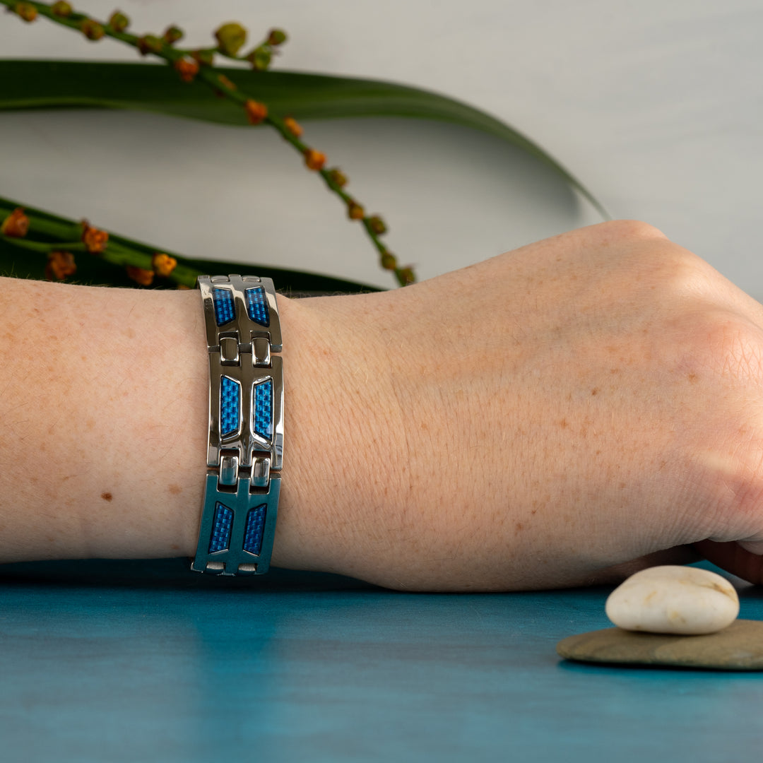 A wrist wears the Magnetic Mobility Yarrow Star 4in1 Magnetic Bracelet, prominently featuring silver links with striking blue carbon fiber accents. The therapeutic piece is showcased against a calming blue backdrop with a blurred natural green plant in the background, emphasizing the bracelet's stylish design and health-oriented features.