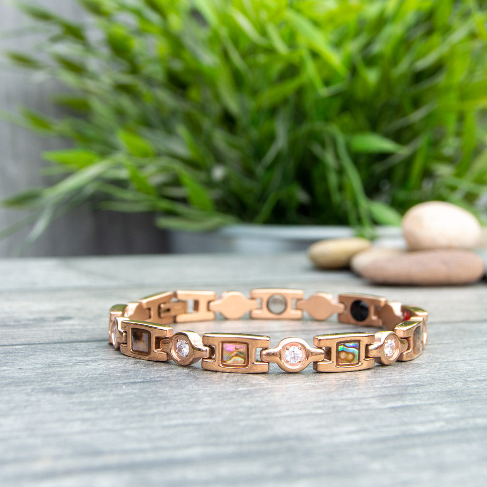 Rose Gold Magnetic Bracelet for Women with Irredessant designs and White crystals. Image shows the bracelet at 45 degree angle showing the 4 health elements with a green plant in the background