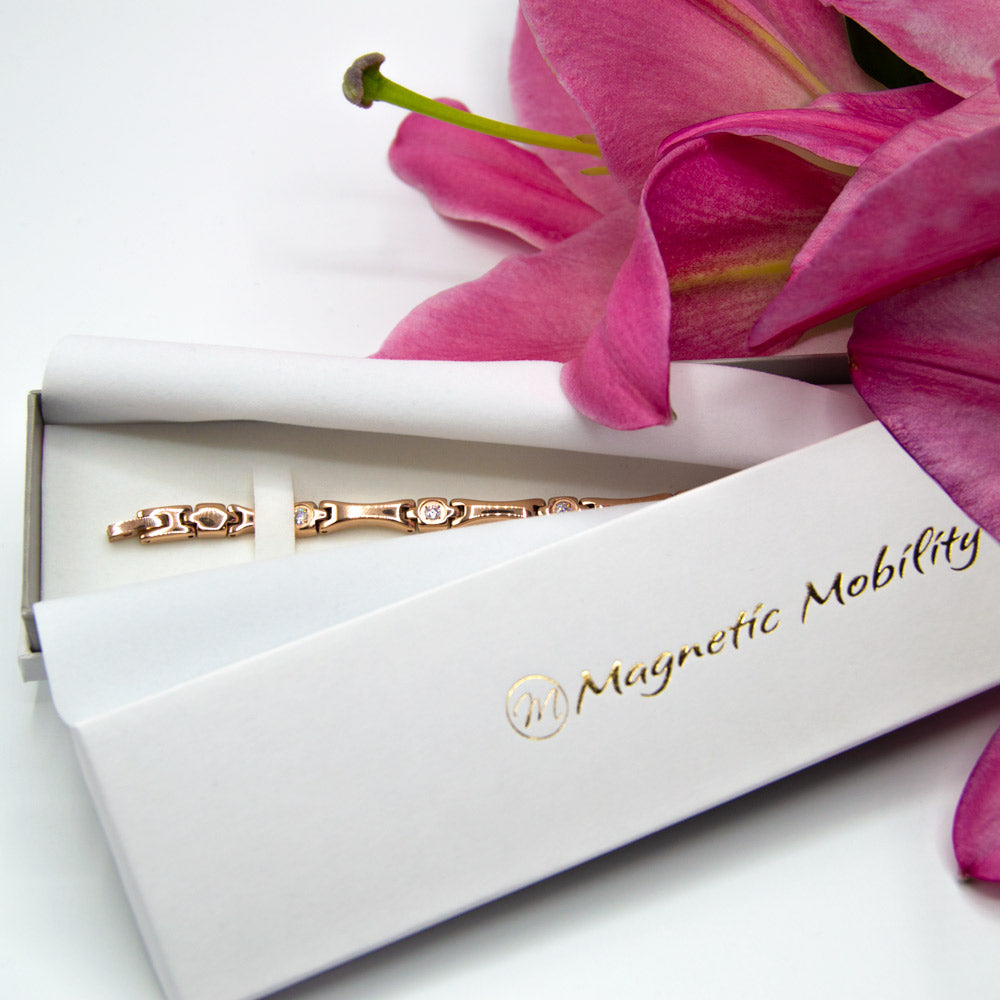 Iris Summer- Womens Magnetic Bracelet in gold with white zirconia stones -top view - inside white jewellery box beside pink lily flower