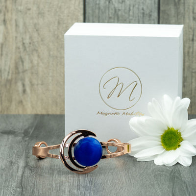 A stylish Orchid Moon Women's Copper Bracelet showcased in front of a plain white square gift box bearing the Magnetic Mobility logo. The bracelet exhibits a stunning crescent moon design with a captivating dark blue stone. Crafted for comfort, the open back allows for a customized fit, offering support for arthritis and back pain. The minimalist white gift box adds a touch of elegance, making it a perfect presentation for this exquisite accessory from Magnetic Mobility.