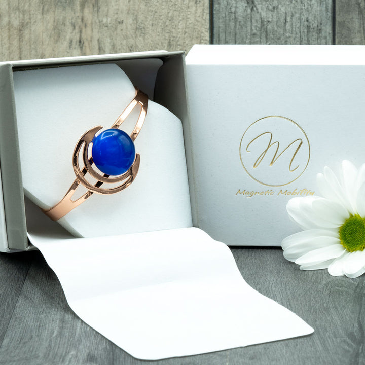 A square luxury eco-friendly gift box, elegantly crafted, showcasing the Orchid Moon Women's Copper Bracelet.  Within the box, the bracelet gleams with sophistication, featuring a crescent moon design with a dark blue stone. Its open back allows for a customized fit, offering support for arthritis and back pain. Experience elevated style and well-being with this exquisite accessory enclosed in the luxurious eco-friendly gift box.